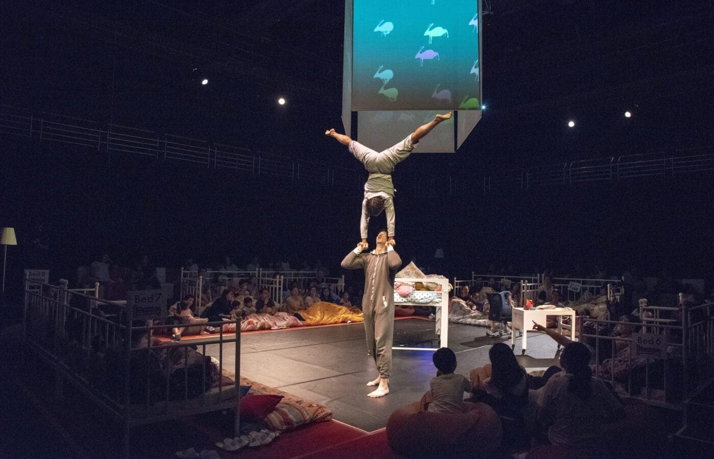 An acrobat lifts another performers into the air upside down. An audience of children watches.