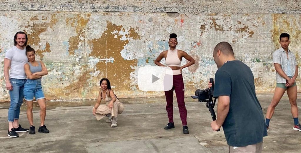 A diverse group of performers stand together smiling whilst being filmed