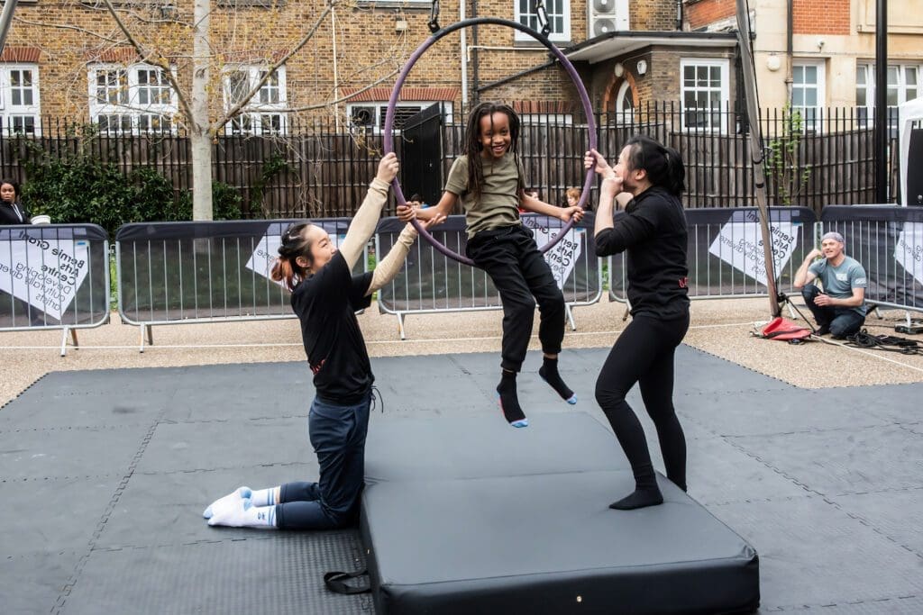 A child sits inside an aerial hoop with a workshop leader either side helping him.