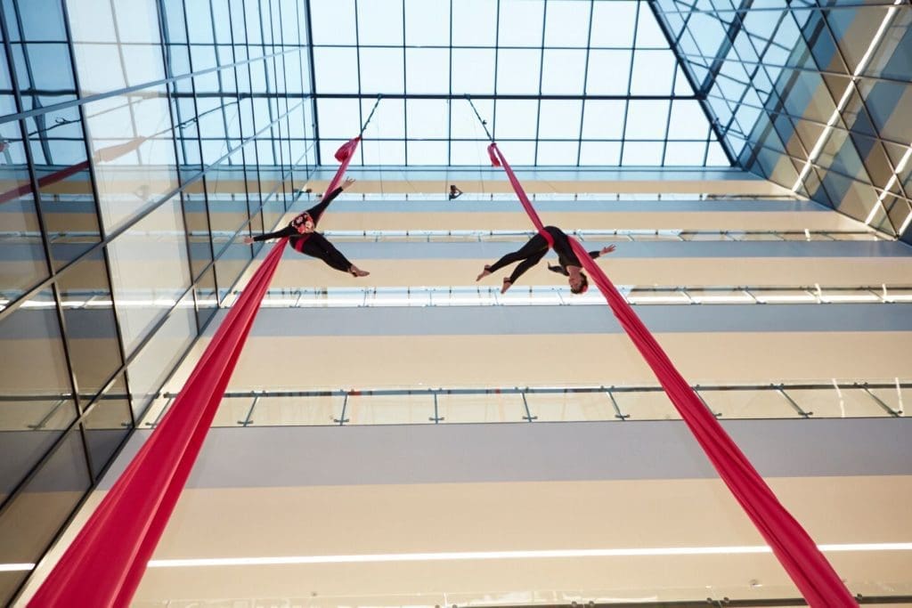 Two aerialists dangle from red aerial ropes from a shopping centre ceiling