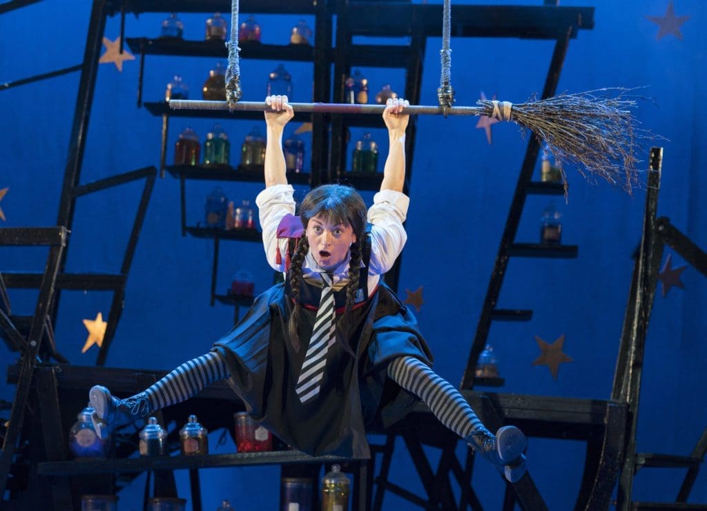 A performer dressed as a witch hangs from a broom-shaped trapeze on a blue stage.