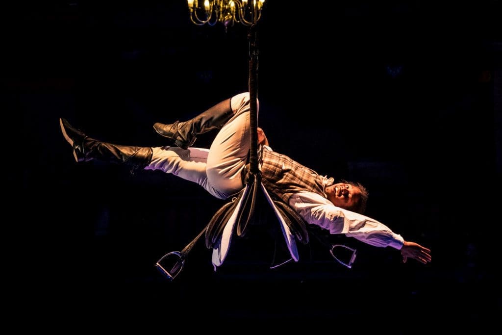 A performer dressed as a jockey lies back across a trapeze with their arms and legs pointed outwards.