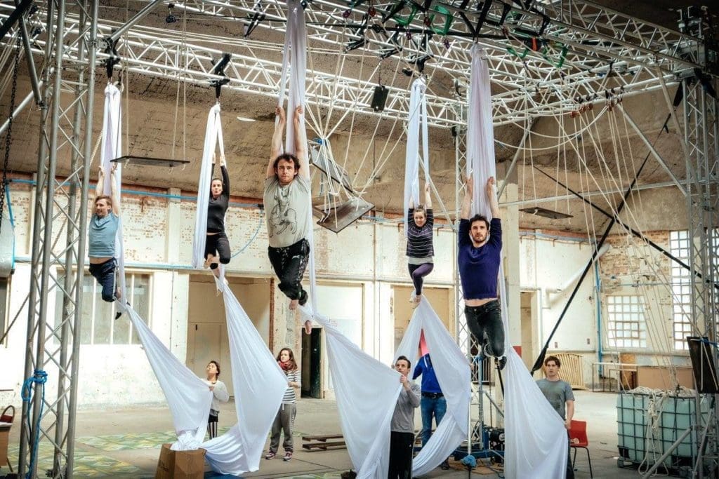 Five aerialists hang from silks while five people on the ground hold the ends of the fabric.