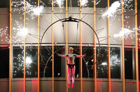 A person is suspended from a pyrotechnics costume that shoots fireworks all around them.