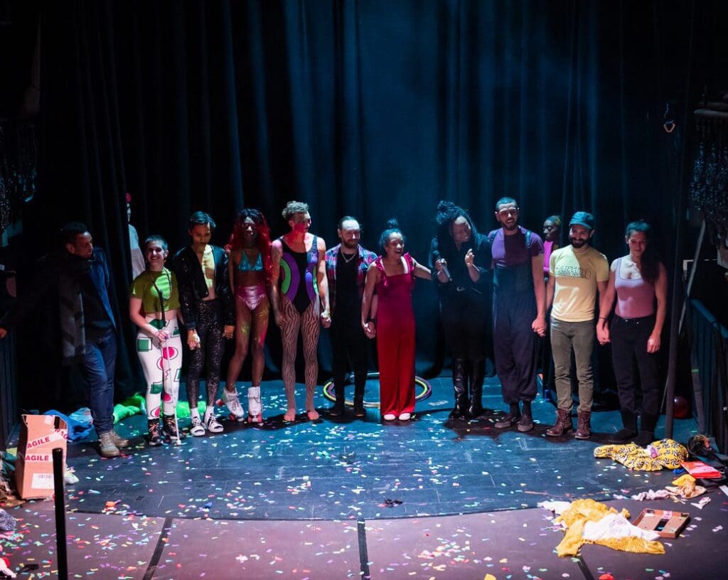 A group of people in different costumes stand holding hands on a dark stage.