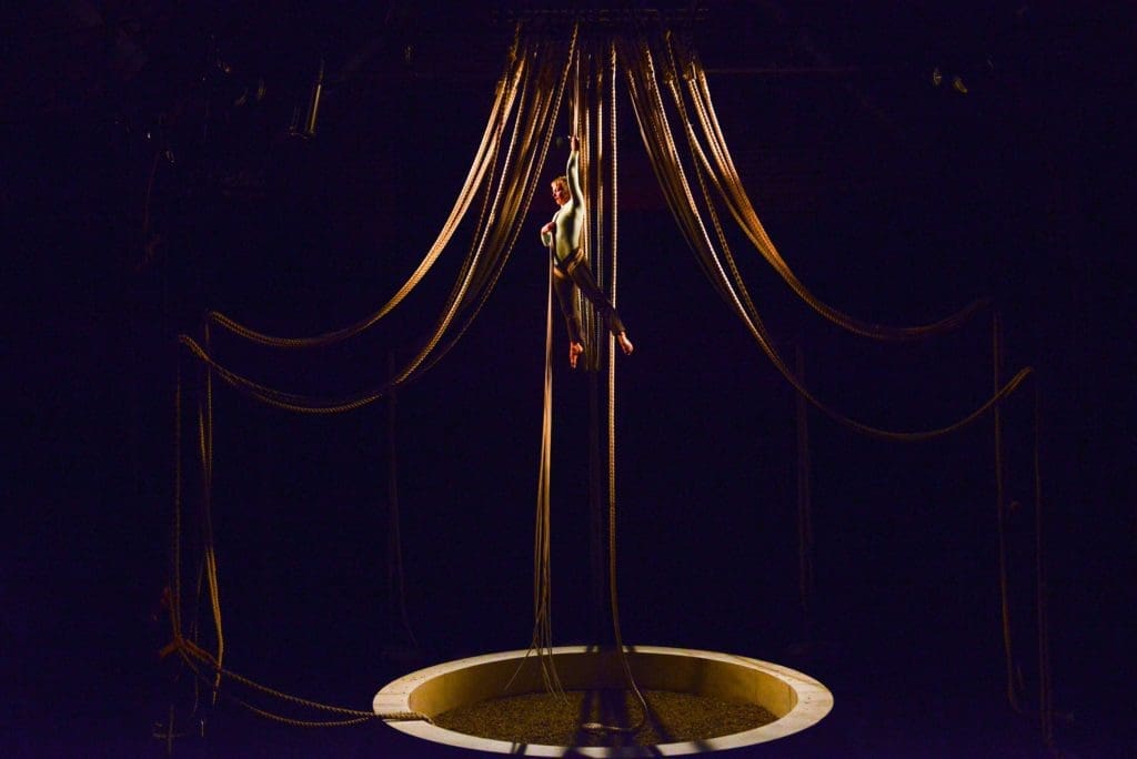 An aerialist dangles in lots of ropes in a dark room.