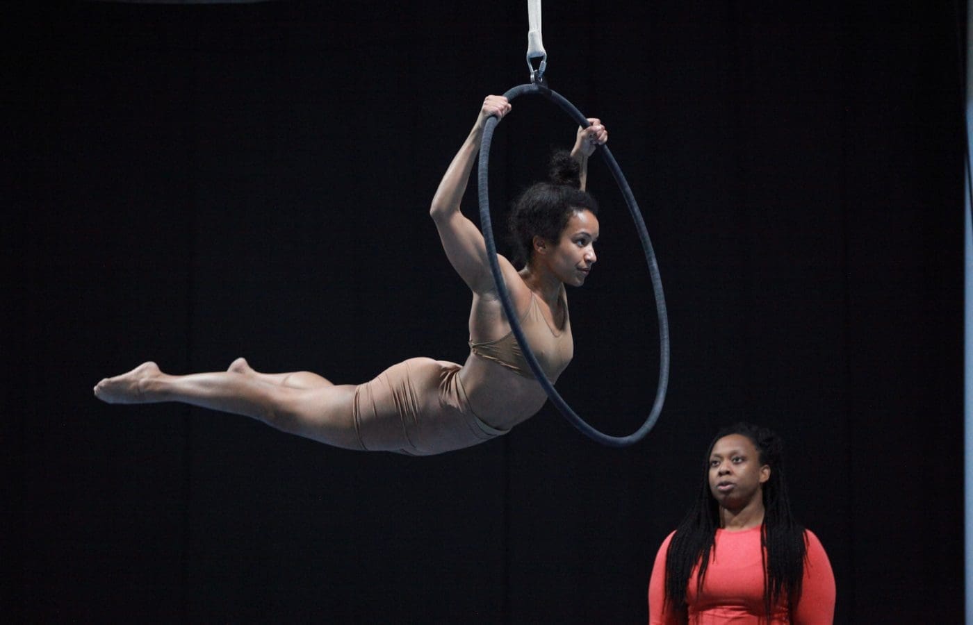 Two women are in a black room. One holds herself up on an aerial hoop while the other stands and watches.