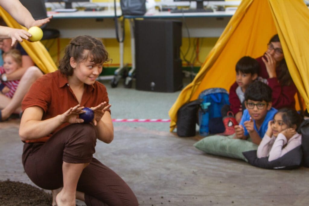 A juggler holds a ball in front of an audience of children sitting in a small tipi