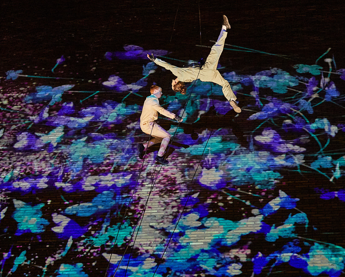 Two people jump whilst suspended from ropes with an abstract projection behind them.