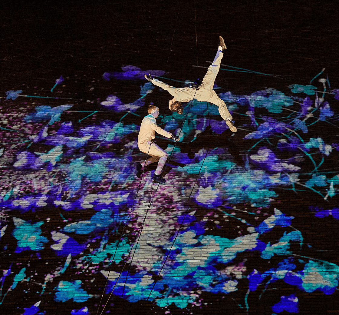 Two people jump whilst suspended from ropes with an abstract projection behind them.