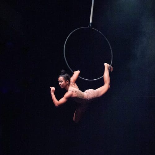A female aerialist hangs from an aerial hoop in a black room