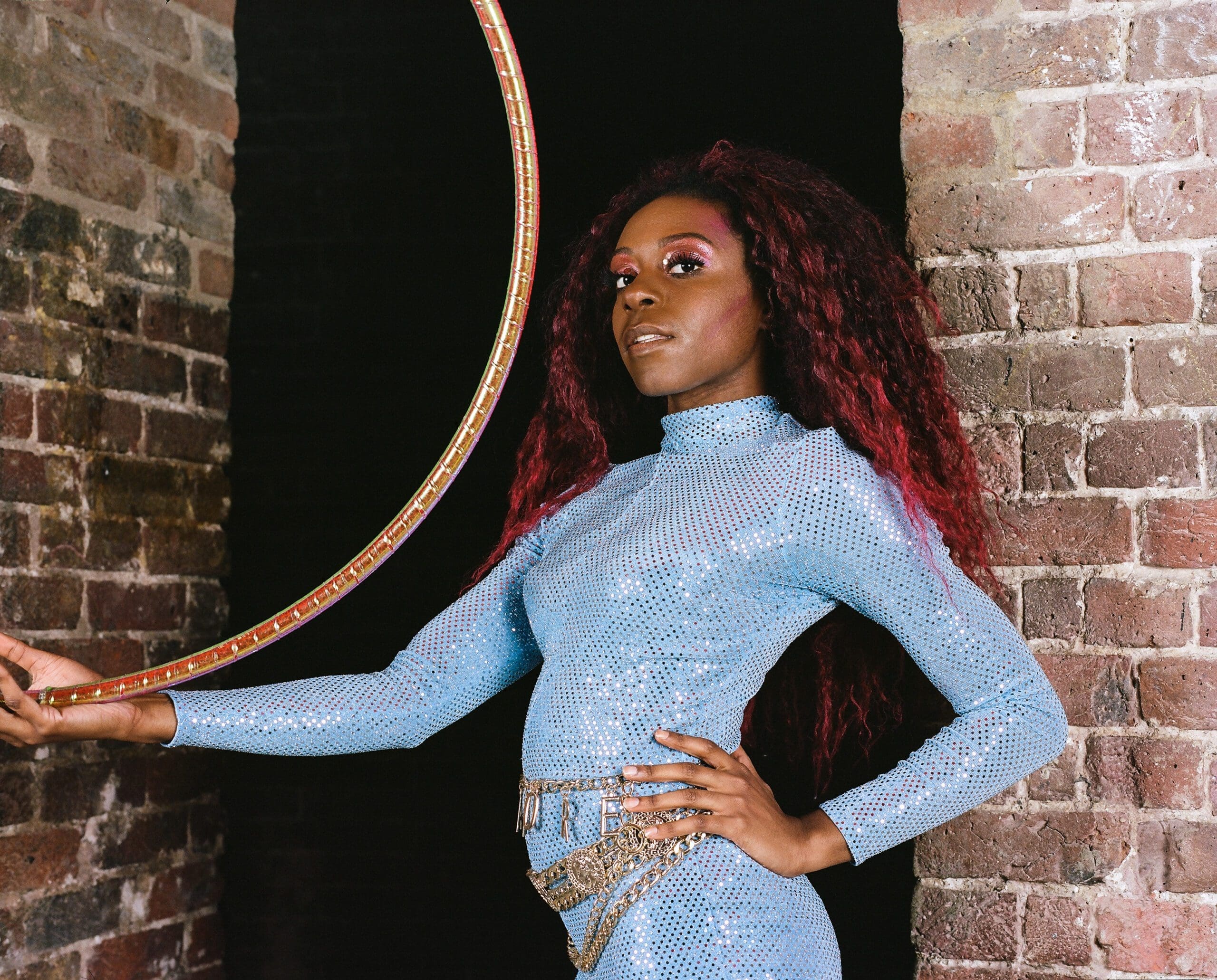 A black woman stands in front of a brick wall in a sparkly blue bodysuit holding a hula hoop