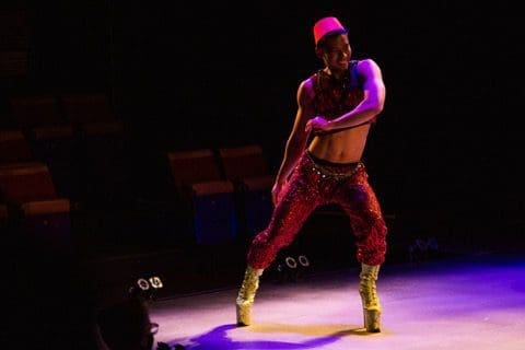 A performer dances and smiles on a purple-lit stage wearing platform heels.