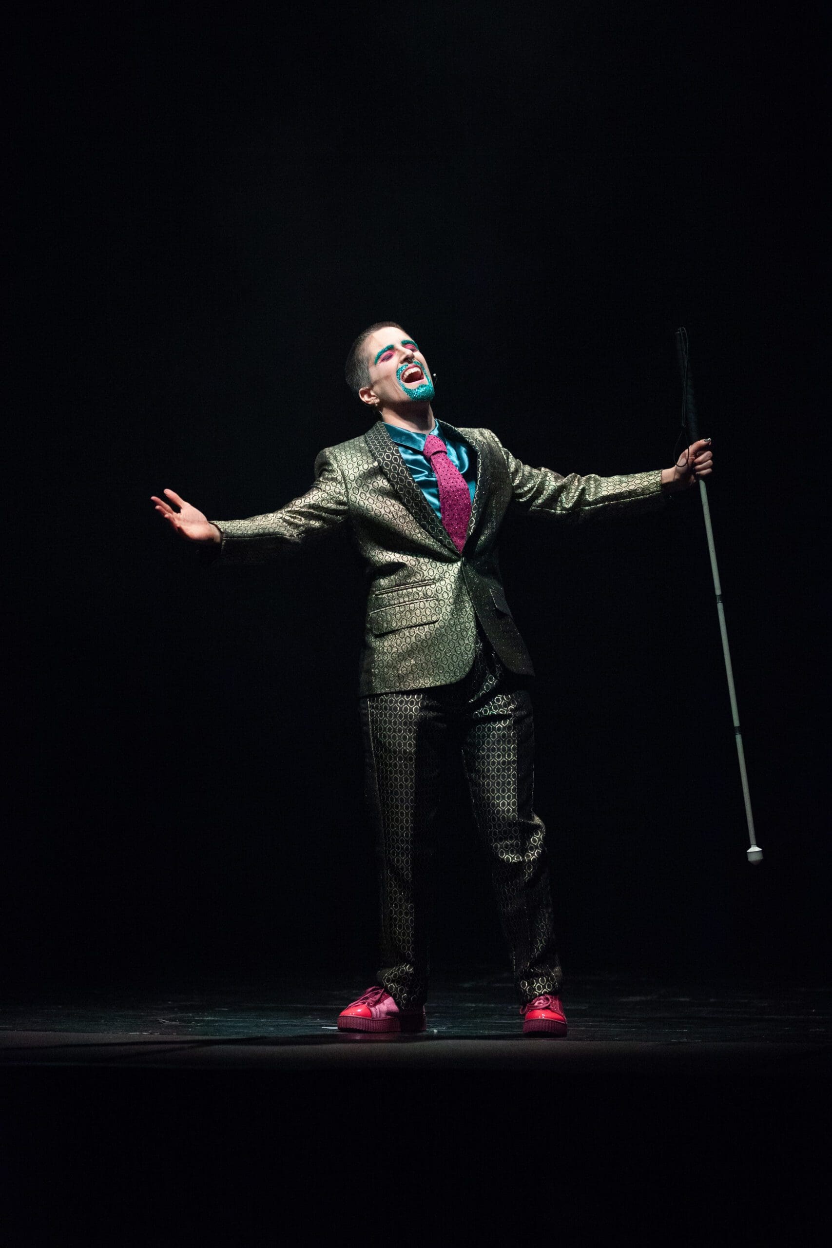 A slim white drag king in a suit and sparkly beard open their arms wide on a dark stage