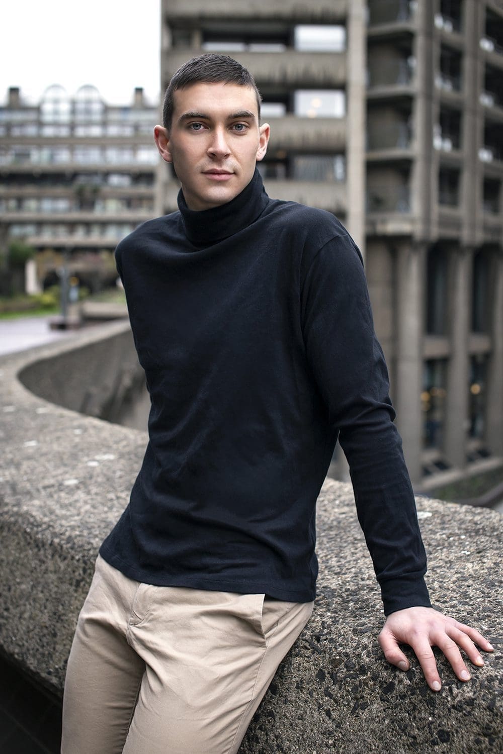 mid-shot of a man leaning on a stone wall outdoors in a navy turtleneck