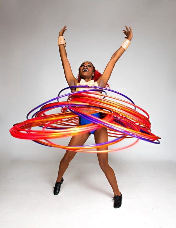 A black woman stands in a white room, spinning lots of hula hoops around her body