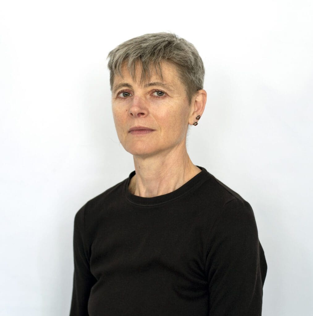 A mid shot of a woman with grey hair standing in front of a white wall in a black top