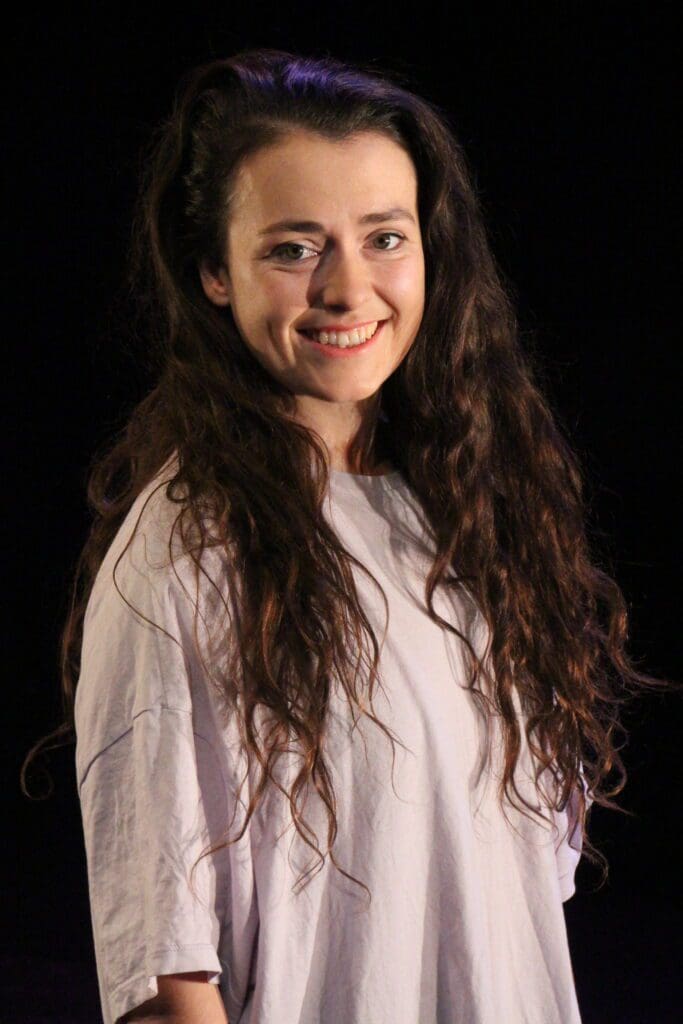 Mid-shot of a 25 year old woman with long brown curly hair who is smiling and wearing a large tshirt