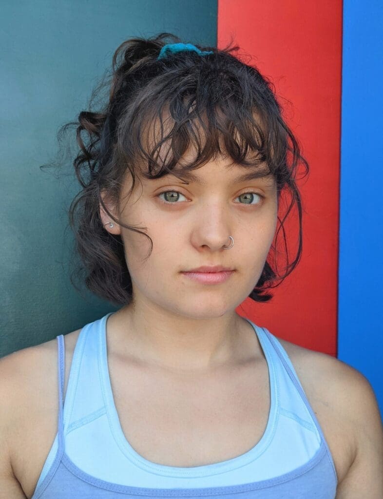 Head and shoulders image of a white 22-year-old with brown curly hair, wearing a light blue tank top. 