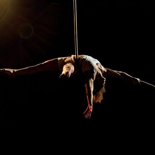 An aerialist dangles backwards from an aerial rope in a dark room. A spotlight shines on them.
