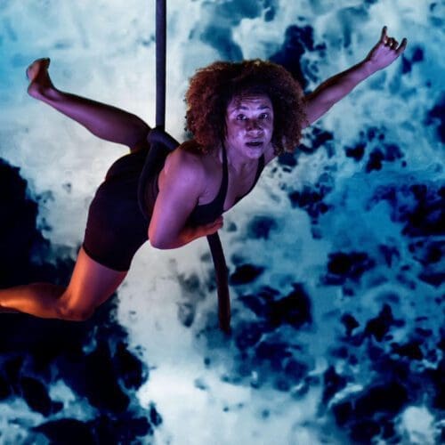 A woman dangles from an aerial rope in front of a blue background