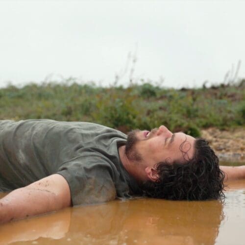 A man lies on his back in a puddle of mud and smiles