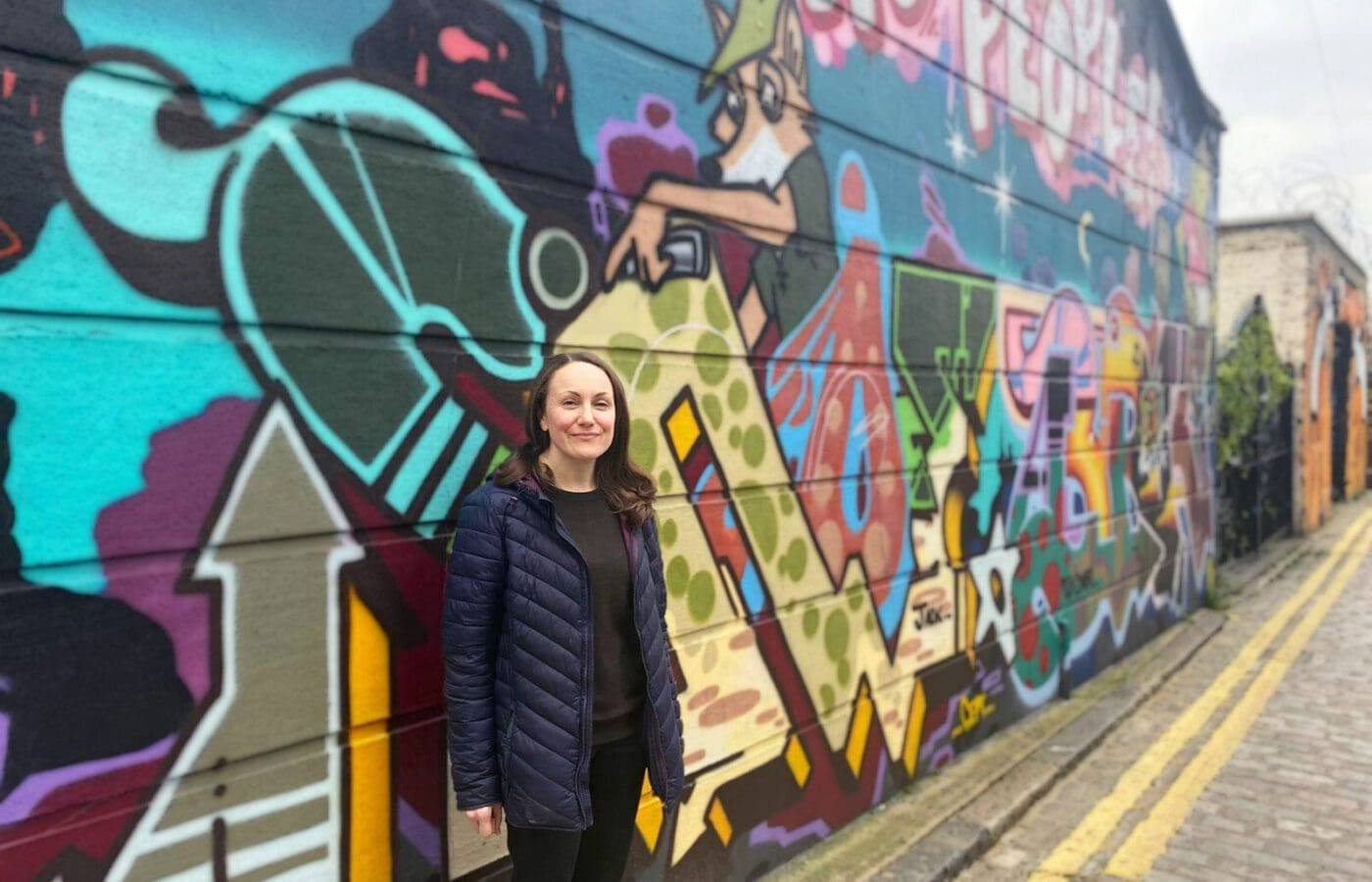 A white woman stands in front of a bright graffiti wall