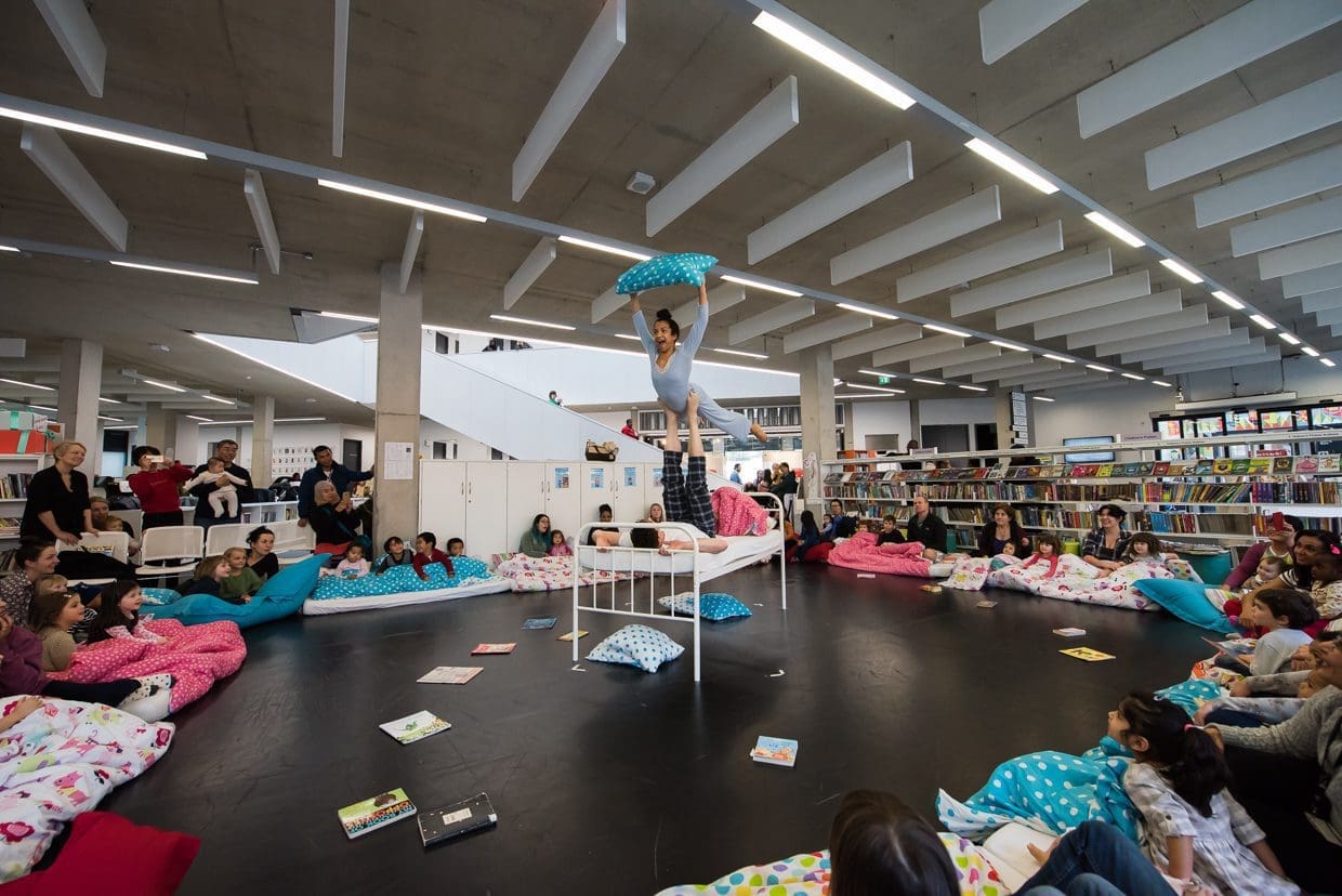 An acrobat balances on another's feet while holding onto a pillow. They are in a library space with an audience of children watching