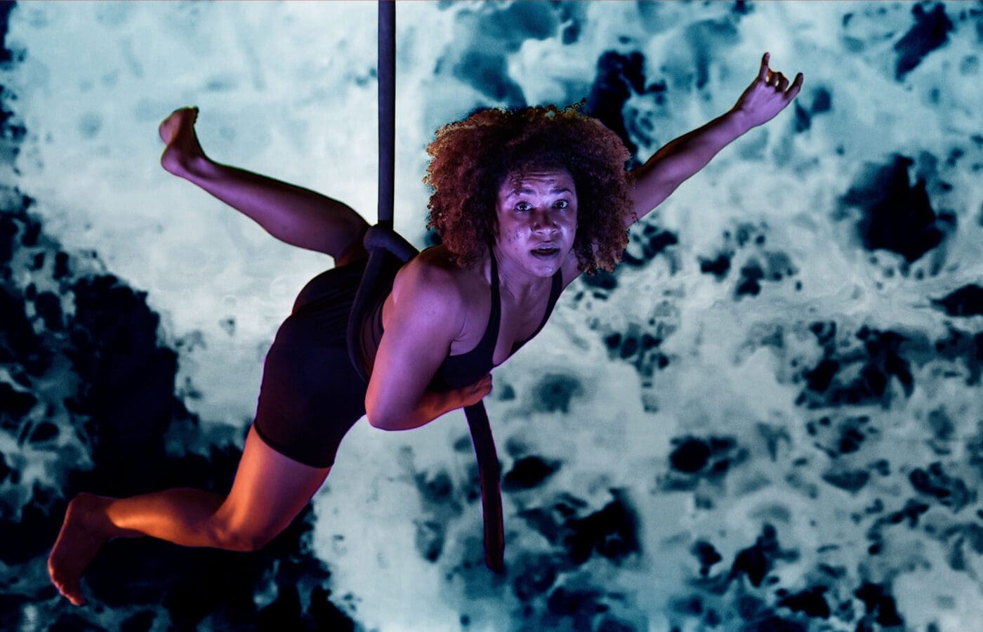 A woman is suspended in the air from an aerial rope. She dangles in front of a mottled blue and black projection