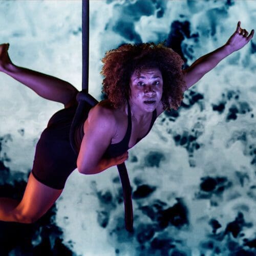 A woman is suspended in the air from an aerial rope. She dangles in front of a mottled blue and black projection