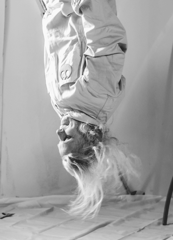 An elderly woman is suspended upside down wearing a space suit. She is a white woman with long white hair