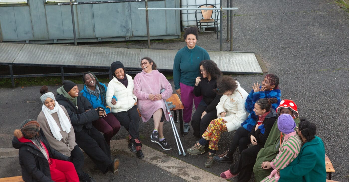 A group of Black women and non-binary people sit outside on chairs in a circle