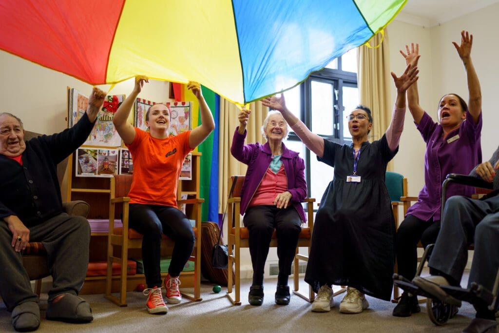 A group of care staff and residents lift a colourful parachute over their heads and laugh