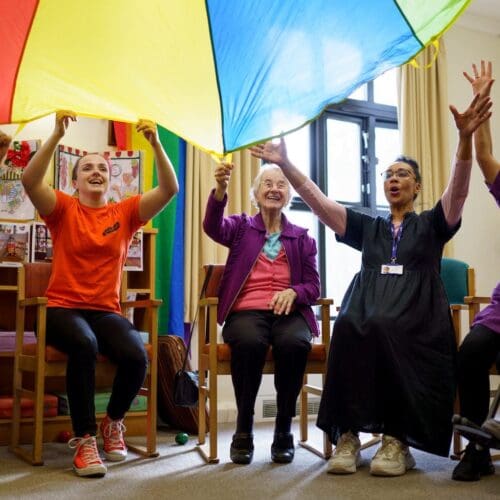 A group of care staff and residents lift a colourful parachute over their heads and laugh