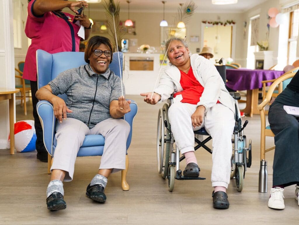 Two older women sit in a care home and laugh as they balance peacock feathers in their hands