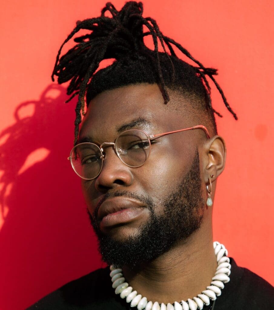 A close up headshot of a man with black dreadlocks and glasses. Wearing a black t-shirt