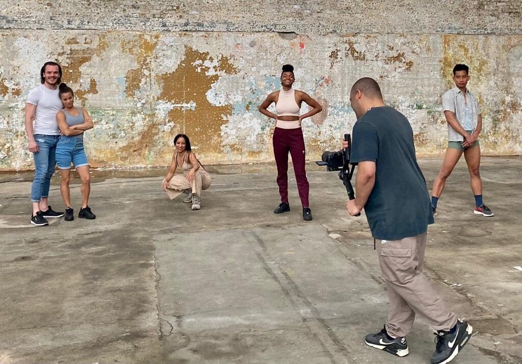 A group of people stand in front of a brick wall and pose as a photographer takes a photo of them,