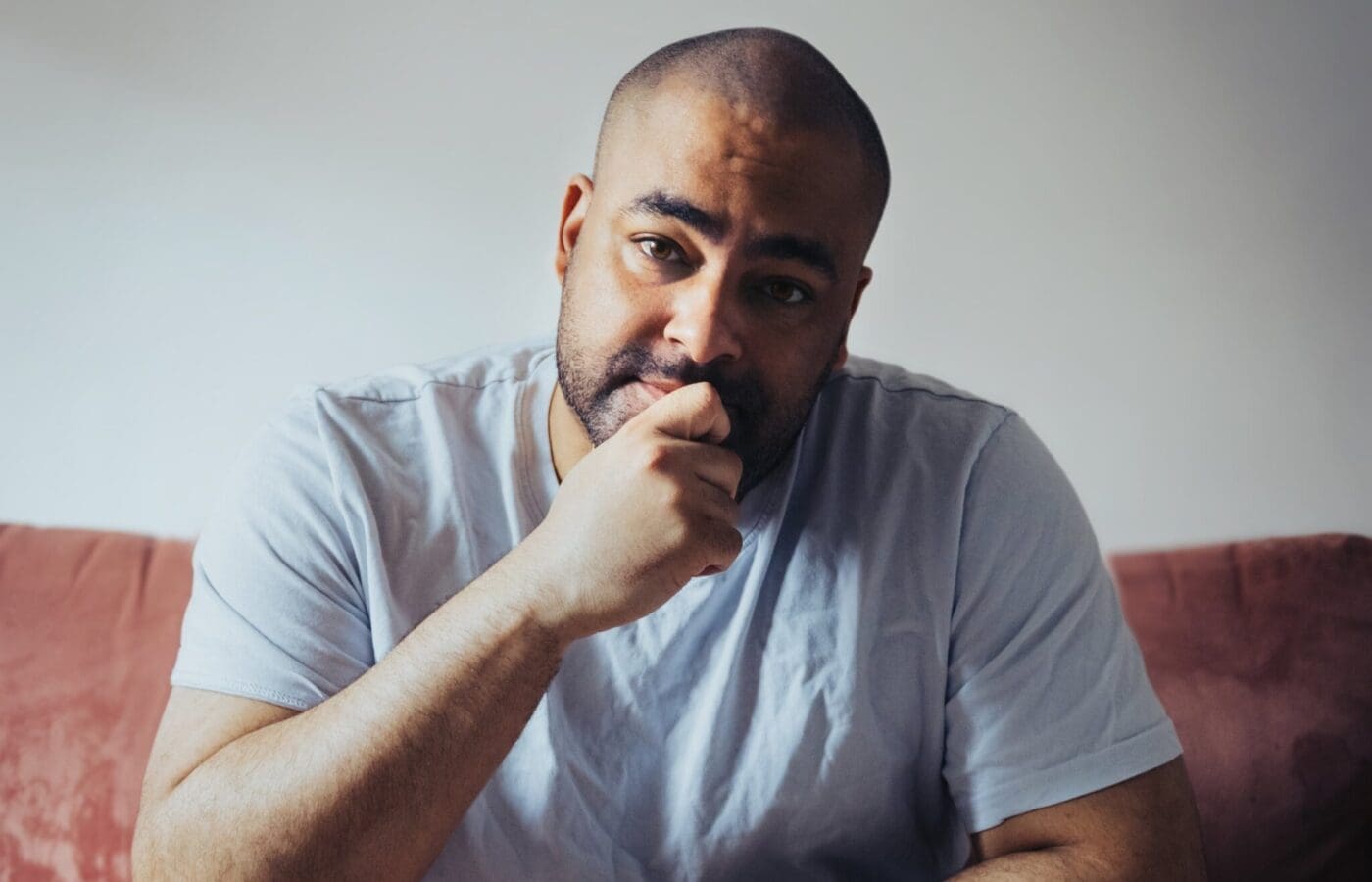 A man sits down with his chin resting in his hand. He has short black hair and a beard. He wears a white tshirt