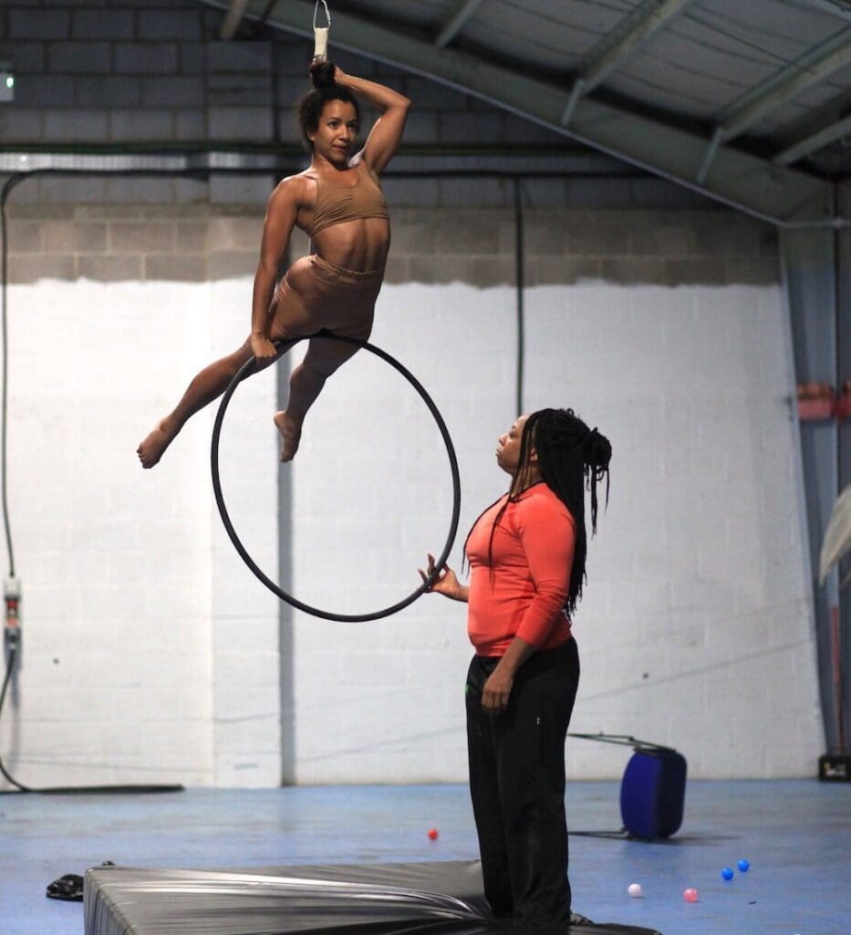 A Black African woman coaches an aerialist as she performs a move on an aerial hoop, as part of Upswing's creative engagement programme.