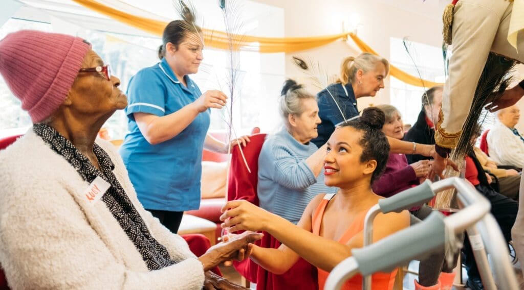 A circus performer helps and elderly lady to balance a feather in her hand in a care home.