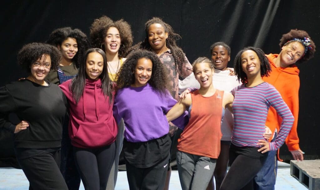 A group of 10 Black women and non-binary circus performers stand in a group in brightly coloured clothes