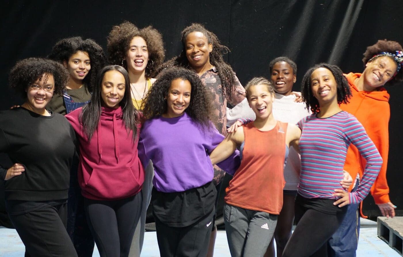 A group of 10 Black women and non-binary circus performers stand in a group in brightly coloured clothes