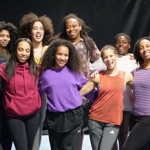 A group of 10 Black women and non-binary circus performers stand in a group in brightly coloured clothes