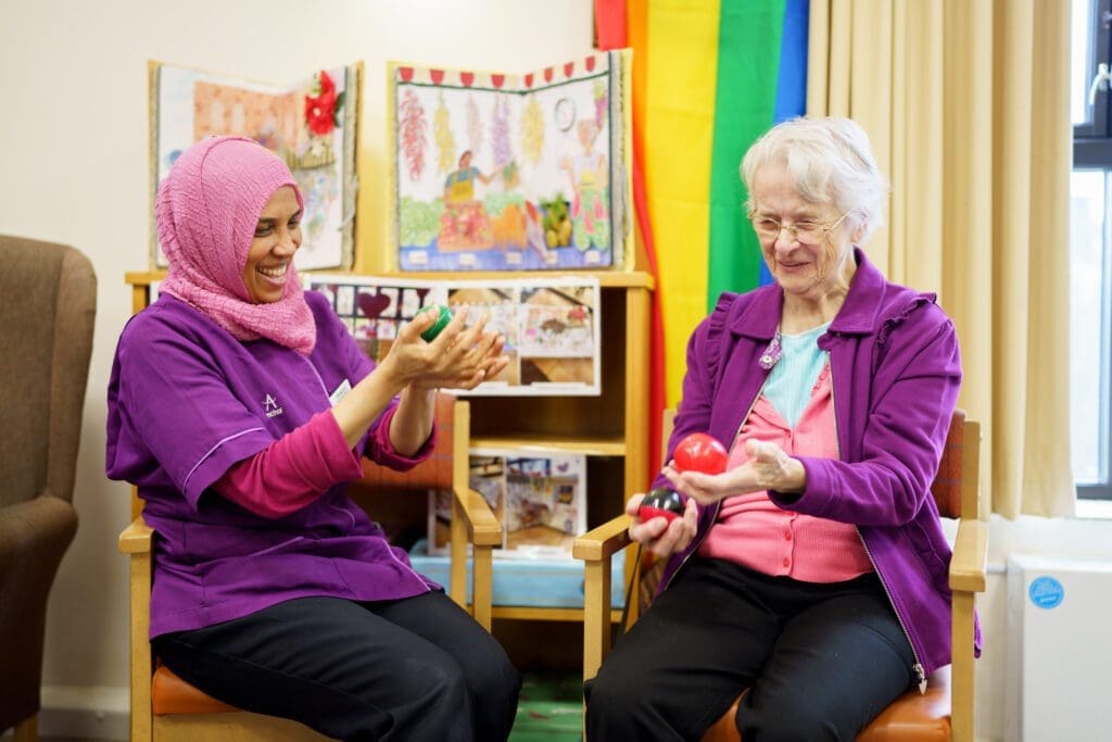 A south asian woman wearing a pink headscarf smiles with an elderly white woman as they both hold juggling balls in a care home setting