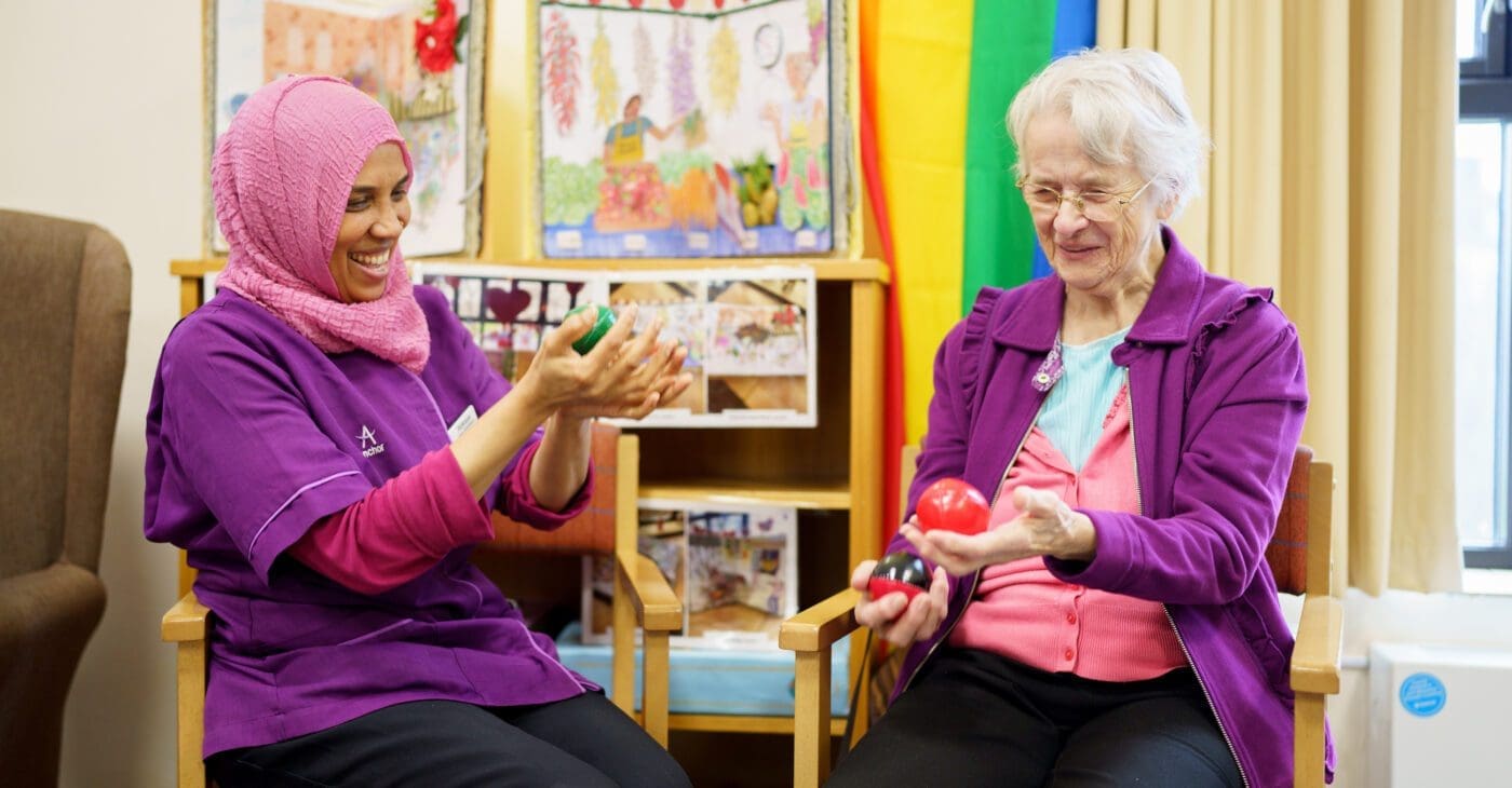 A south asian woman wearing a pink headscarf smiles with an elderly white woman as they both hold juggling balls in a care home setting