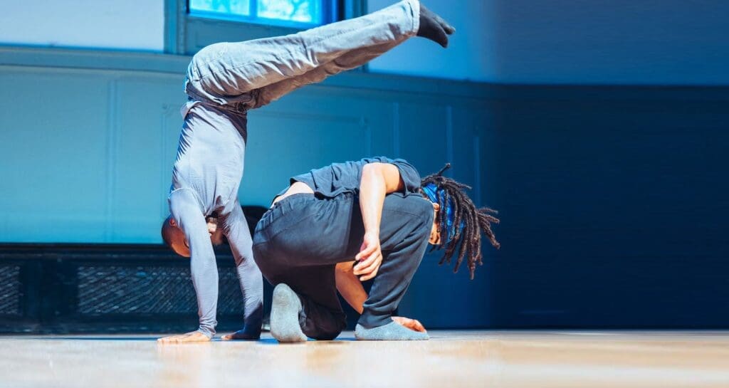 An black male acrobat jumps and does a handstand over the top of another