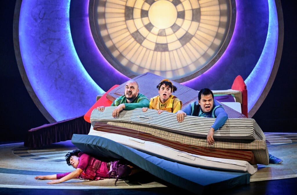 A group of acrobats sit on top of mattresses squashing someone underneath
