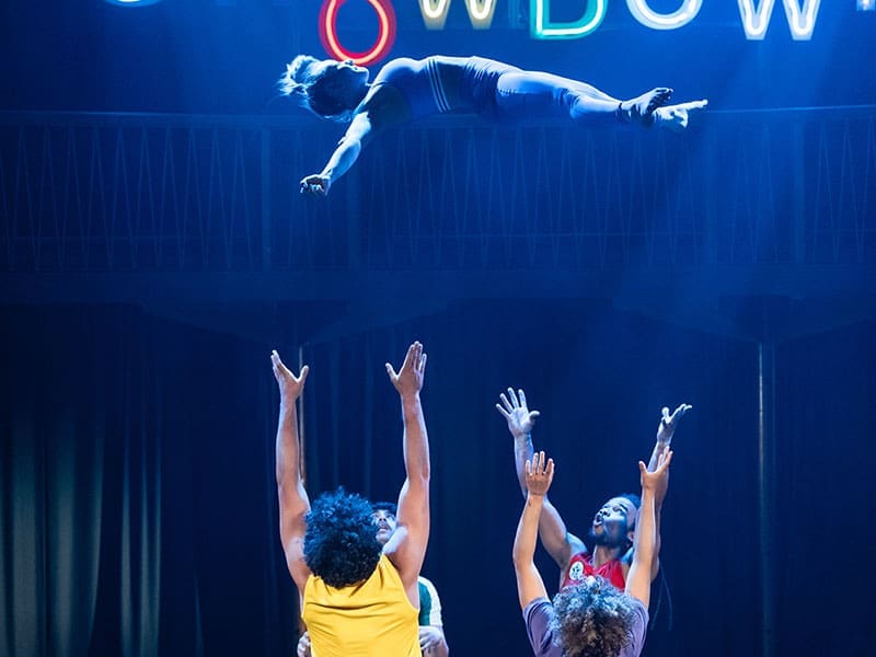 A group of acrobats throw someone high into the air on a darkly lit stage