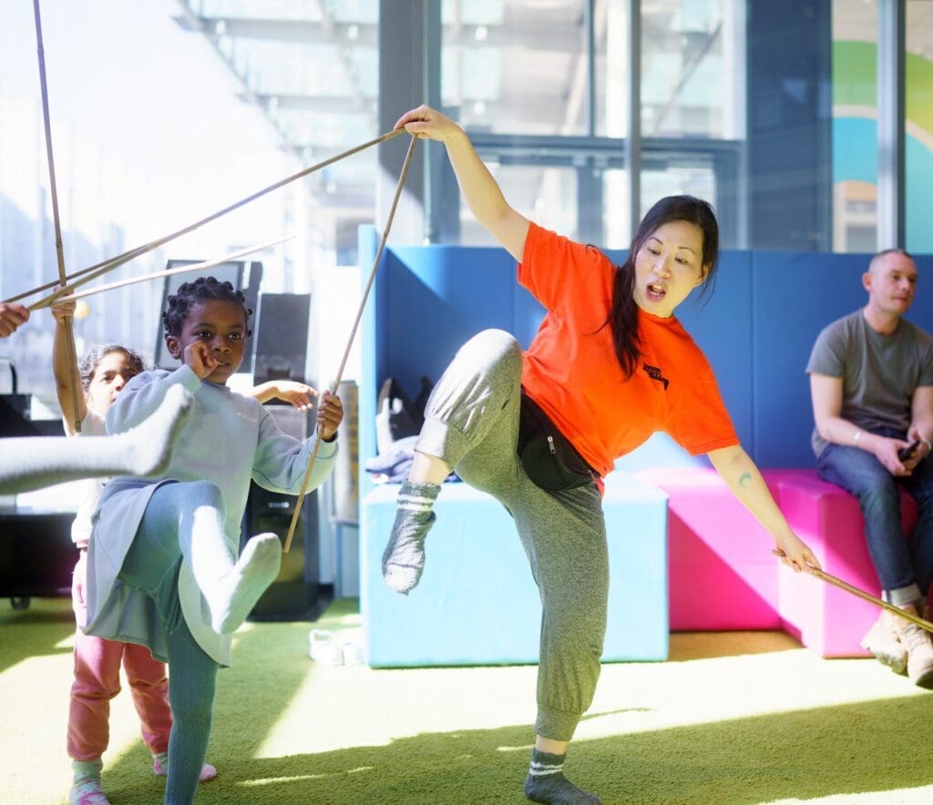 A woman runs a circus workshop and holds some sticks and dances with some children