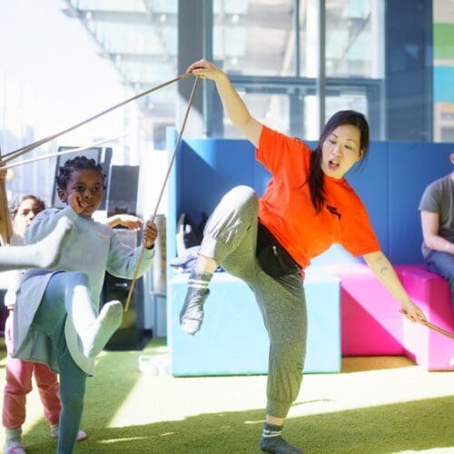 A woman runs a circus workshop and holds some sticks and dances with some children